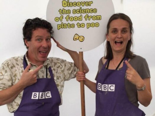 Jon and Sarah Bearchell stand together holding a lollypop sign reading 'From Plate to Poo'.
